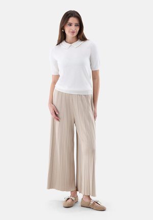 Frau trägt ein weißes Kurzarmtop mit detailliertem Kragen, beige weite plissierte Hosen und beige Ballerinas mit Schleifen.