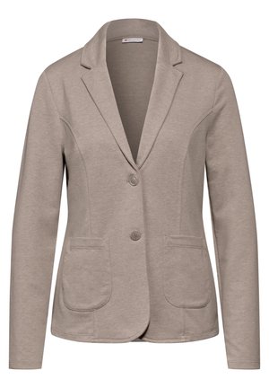 Persoon draagt een beige blazer over een wit shirt met blauwe spijkerbroek, één hand in de zak en de andere ontspannen naast het lichaam.