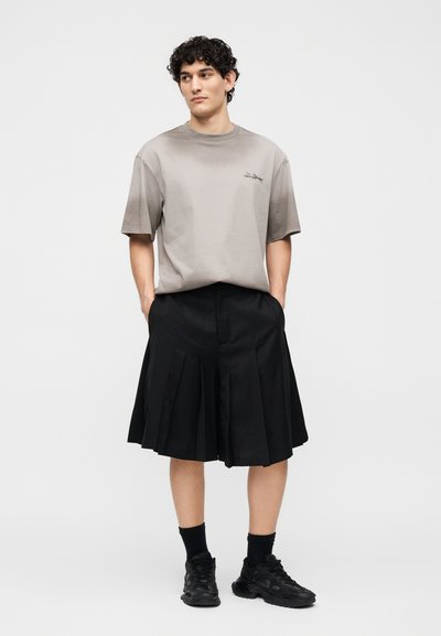 Νέος άνδρας στέκεται με τα χέρια στις τσέπες, φοράει ένα φαρδύ γκρι T-shirt, μαύρη πλισέ φούστα μέχρι το γόνατο, μαύρες κάλτσες και μαύρα αθλητικά παπούτσια.