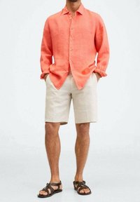 Camicia di lino corallo con bottoni, abbinata a pantaloni corti beige chiaro. Sandali marroni con cinturini completano l'outfit, mettendo in mostra uno stile estivo rilassato.