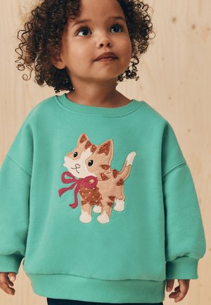 Nuori lapsi, jolla on kiharat hiukset, pukeutuu turkoosiin collegepaitaan, jossa on ommeltu kissanpentu vaaleanpunainen rusetti päässään, seisoo vaaleanpuun taustaa vasten.