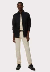 Schwarzes Hemd mit Knöpfen, beigefarbene, eng anliegende Hose, schwarze Schuhe, mit einem hellfarbenen Unterhemd und einem Gürtel. Glatter Stoff und maßgeschneiderte Passform.