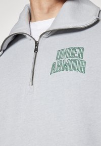 Sudadera gris con cremallera, hecha de una tela suave, que cuenta con un cuello alzado y un logo bordado en verde de "UNDER ARMOUR" en el pecho.