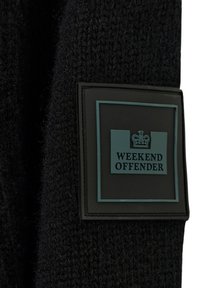 Schwarze Strickmütze aus strukturiertem Stoff. Mit einem rechteckigen Aufnäher in Schwarz und Türkis, auf dem "WEEKEND OFFENDER" steht.