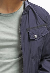 Navyblauwe lichtgewicht jas met een ritssluiting, met een borstzak met een drukknoop en een lichte glans, gedragen over een grijs T-shirt.