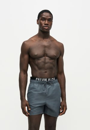 Homme musclé debout sur fond blanc, portant un short de bain gris avec la ceinture Calvin Klein visible, regardant droit devant.