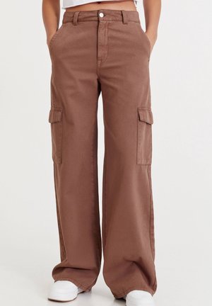 Brązowe, szerokie spodnie typu cargo z przednimi kieszeniami oraz kieszeniami bocznymi z klapami, noszone z białymi sportowymi butami i krótkim topem na jednolitym tle.