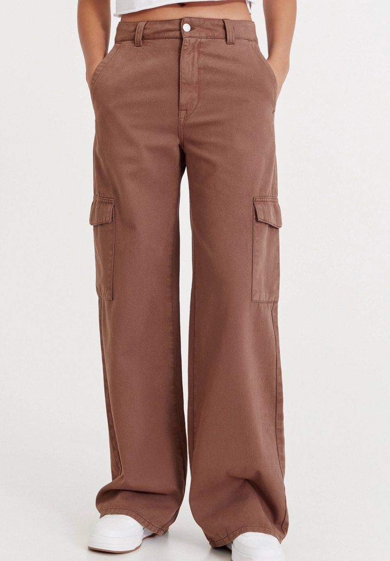 Pantalon cargo marron à jambes larges avec poches avant et poches latérales à rabat, porté avec des baskets blanches et un top court sur un fond uni.