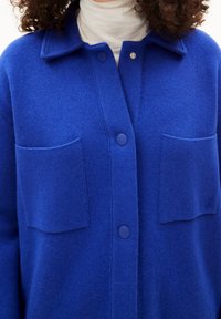 Blå stickad kofta med en strukturerad yta, med krage, framknäppning med knappar och två fickor. Slät tyg, avslappnad design.