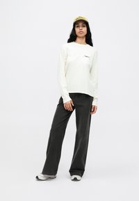 Roomkleurig shirt met lange mouwen waarop "OBEY" in het zwart gedrukt staat, gecombineerd met donkergrijze wijde broekspijpen en een gele pet. De schoenen zijn wit en grijs.