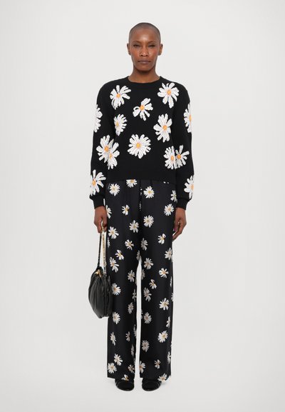 Schwarzer Pullover mit weißer und gelber Gänseblümchen-Stickerei, kombiniert mit weiten schwarzen Hosen, die das gleiche Blumenmuster aufweisen. Schwarze Tasche, schlichte Schuhe.