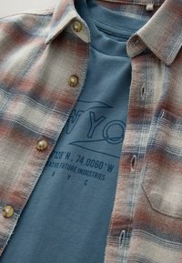 Cămașă din flanel în carouri crema, roșu și albastru, cu butoane rotunde. Sub aceasta, un tricou albastru cu text grafic și coordonate.