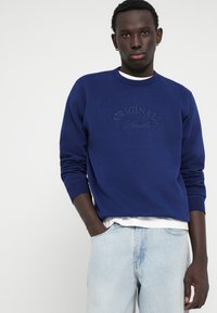 Sweatshirt bleu marine avec des poignets et un col côtelés. Logo "ORIGINALS" brodé dans une teinte plus claire. Superposé à une chemise blanche, associé à un jean clair.