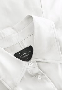 Blusa bianca con colletto classico, chiusura a bottoni e texture setosa. Etichetta che recita "Jascha, Stoccolma," taglia S.