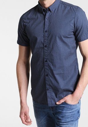 Mann trägt kurzärmliges dunkelblaues Hemd mit kleinem weißem Kreis-Muster und blaue Jeans, Hand in der Hosentasche.