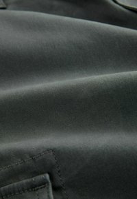 Dunkelgrüner Stoff mit einer glatten Textur, der dezente diagonale Linien und eine genähte Nahtdetail am Rand aufweist.