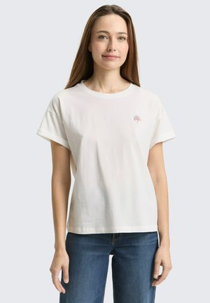 Frau mit langen braunen Haaren, die ein weißes T-Shirt mit kurzen Ärmeln trägt, auf der Brust ein kleines, buntes Vogel-Logo und blaue Jeans.