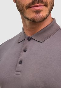 Szara koszulka polo o fakturowanym wykończeniu, z kołnierzykiem i trzema czarnymi guzikami. Widoczne są zbliżone szczegóły tkaniny i kolorystyki.