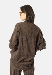 Brun, myk jakke i semsket stoff med avslappet passform, oppbrettede ermer og bakre sømsdetalj, båret over matchende bukser mot en ensfarget bakgrunn.