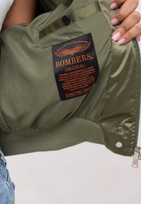 Veste bomber verte à finition satinée, avec des poignets côtelés et une étiquette orange et noire comportant du texte et un graphisme d'aigle à l'intérieur.