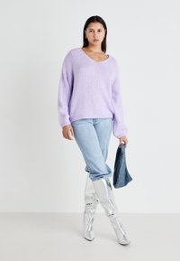 Lila gebreide trui met V-hals, gecombineerd met lichtblauwe jeans. Model draagt glanzende zilveren kniehoogte laarzen en houdt een denim jack vast.