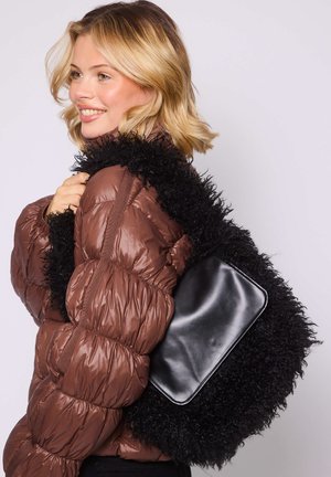 Glimlachende vrouw met kort blond haar draagt een bruine donzen jas en heeft een grote zwarte, zachte handtas met een glad leren stuk op de schouder.