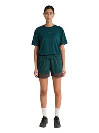 Kvinde stående iført en mørkegrøn løs T-shirt og matchende shorts med hvide sokker og sorte sportssko på hvid baggrund.