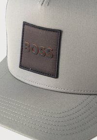 Casquette de baseball gris clair avec un patch carré marron foncé affichant le mot "BOSS" en lettres noires en relief avec un contour orange.