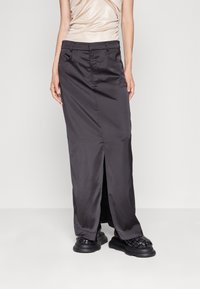 Kvinna klädd i en svart lång kjol med slits framtill, beige rynkad topp och svarta flätade plattformsskor stående mot en enfärgad bakgrund.