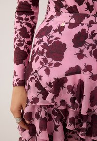 Roze jurk met bordeauxrode bloemenpatroon, met lange mouwen en een gerimpelde zoom. Kleine gouden hardwareaccenten bij de taille.