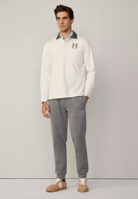 Beige långärmad tröja med krage och "H"-logotyp, kombinerad med grå sweatpants med ett "H"-emblem och beiga sneakers.