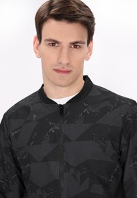 Giubbotto bomber nero con motivo geometrico, colletto alto e cerniera frontale. Realizzato in tessuto liscio e leggermente testurizzato.