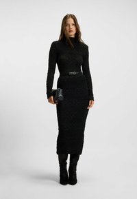 Schwarzes langärmliges Kleid mit strukturiertem Muster, tailliertem Oberteil und Midilänge-Rock, kombiniert mit einem schwarzen Gürtel und einer kleinen Handtasche.