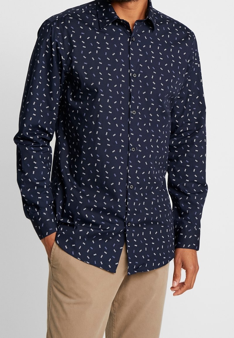 Homme portant une chemise bleu marine à boutons avec de petits motifs blancs en forme de clé et un pantalon beige, debout avec une main dans la poche.