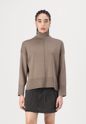 Suéter marrón de punto con cuello alto y puños y dobladillo acanalados; presenta un corte recto y un sutil detalle de costura. Llevado sobre una falda negra.
