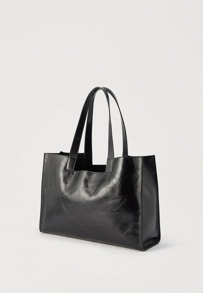 Μαύρη ορθογώνια δερμάτινη τσάντα χειρός με δύο μακριές λαβές, εμφανίζεται να στέκεται όρθια σε ανοιχτό φόντο.