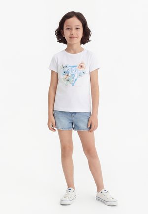 Rövid sötét hajjal rendelkező fiatal lány, fehér pólót visel, mely pillangó- és virágmintás, farmer rövidnadrágot és fehér tornacipőt, fehér háttér előtt áll.