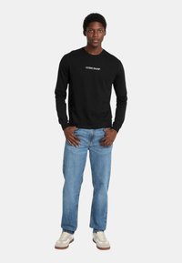 Maglietta nera a maniche lunghe con logo "GUESS JEANS", abbinata a jeans in denim blu chiaro e sneakers beige con accenti bianchi. Stile e vestibilità casual.