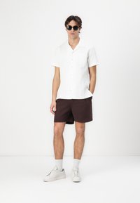 Camisa de botones de manga corta blanca con una textura sutil, pantalones cortos marrones, calcetines grises y zapatillas blancas, con gafas de sol oscuras.