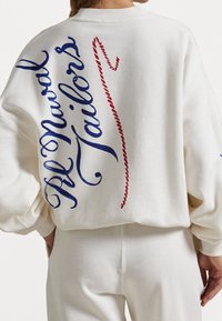 Vit sweatshirt med blå kursivtext "Al Naval" och röda kursivaccenter. Lång lös passform med sänkta axlar och mjuk bomullstextur.