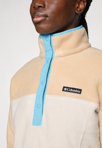 Beige en crème fleecetrui met een hoge kraag, blauwe drukknoopjes en een zwart Columbia-logo op de borst. Zachte textuur, casual ontwerp.