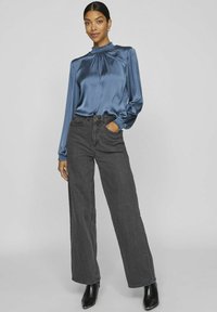 Blusa blu in satin con collo alto e dettagli arricciati, abbinata a jeans grigi scuri a vita alta e gamba larga; stivaletti neri alla caviglia.