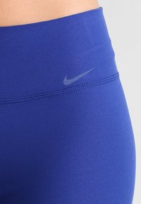Modré legíny s vysokým pasem vyrobené z elastického materiálu. Vlastnosti zahrnují malé šedé logo Nike na pase. Hladký, těsně padnoucí design.