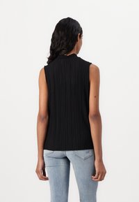 Kobieta z ciemnymi falującymi włosami, ubrana w bezrękawnik z czarnego prążkowanego materiału i jasnoniebieskie dżinsy, stoi twarzą do białego tła.