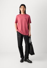 Tricou roz din bumbac cu logo imprimat, asortat cu blugi din denim închis. Modelul ține o geantă neagră și poartă cizme negre, stând pe un fundal neutru.