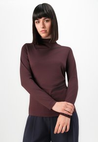 Vrouw met steil donker haar, gekleed in een lange mouwen bordeauxkleurige top en donkere broek, staand tegen een eenvoudige witte achtergrond.