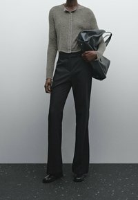 Henkilö, joka on pukeutunut harmaaseen nappikauluspaitaan, mustiin korkeavyötäröisiin leveälahkeisiin housuihin, mustiin kenkiin ja pitää suurta mustaa nahkalaukkua käsivarrellaan.