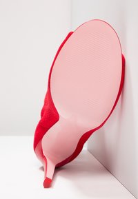 Scarpa con tacco alto in suede rosso, con punta affusolata e suola in gomma rosa liscia. Il tacco è sottile e la scarpa presenta una superficie testurizzata.