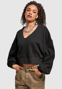 Ungdommelig kvinne med krøllete hår som har på seg en svart kortermet cardigan med knapper, en tykk sølvkjede, og beige høyhøydde bukser.