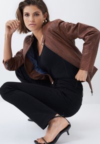 Chaqueta de cuero marrón con cierre de cremallera y diseño acolchado, combinada con una blusa negra, pantalones negros y tacones negros. Modelo en una pose en cuclillas.